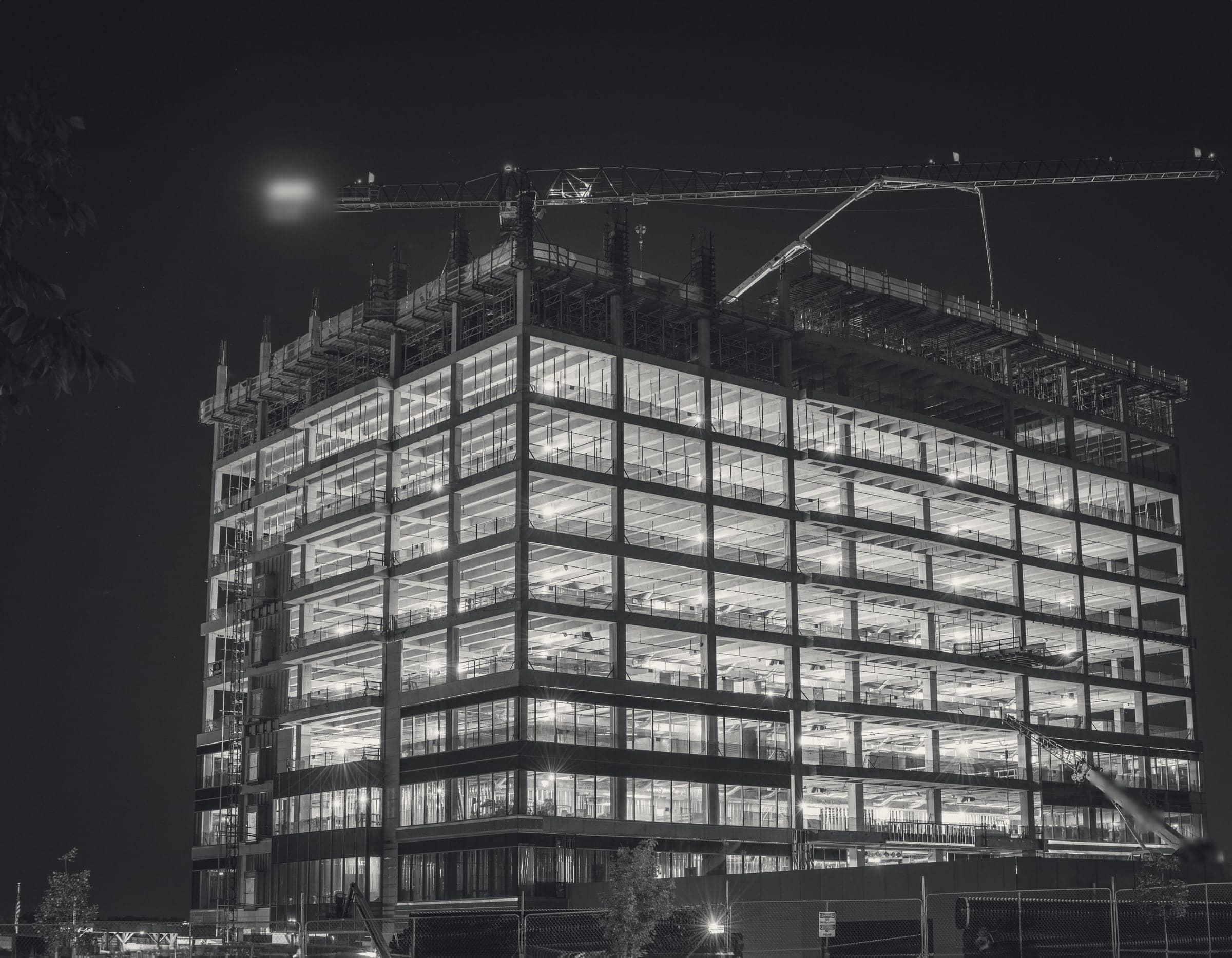 A concrete-framed building under construction at night, every floor lit, with a tower crane across the top.